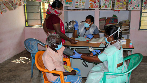 In a clinic in India, an elderly man is seated across from a health care provider and is having his blood pressure read by a woman in a turquoise sari. Behind them is a desk where another provider is seated and speaking to a woman standing. They are reviewing a document together.