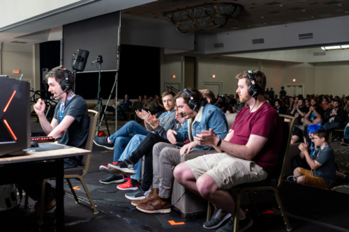 A man sits at a table facing a gaming console and wearing a headset. Four other adults sit behind him on a couch also wearing headsets. All are on a stage in a hotel ballroom. There is a large crowd of people behind them watching the game being projected on a screen that is not visible in the photo.