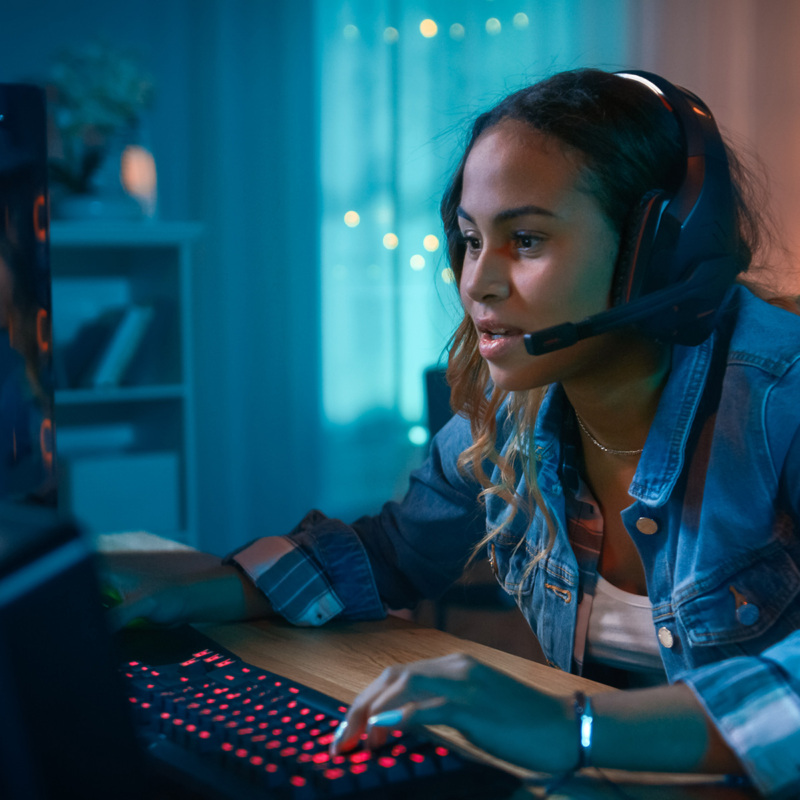 A teen girl sits at a desk facing a computer screen with both hands on the keyboard. She is wearing gaming headphones, leaning in and staring intently at the screen.