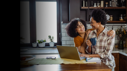 Two black women in their 40s stand close together in a kitchen while gazing at each other with smiles. The woman on teh right is holding a coffee mug and has a computer in front her. The woman on the left is embracing the other woman from behind with both her hands on the other woman's shoulder.