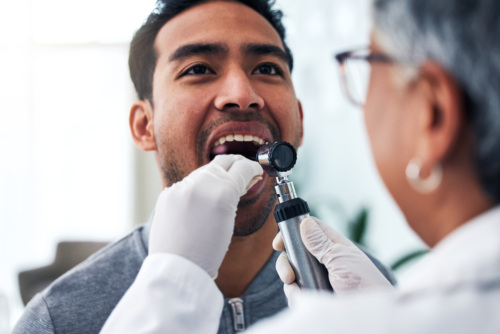 A closeup of a man in his 30s getting an oral exam. A health care provider is facing the man and slightly out of focus. She is holding a tongue depressor in the man’s mouth and holding a magnifying scope with the other hand.