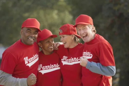 Four adults in their 50s dressed in red baseball jerseys and caps. There is a Black man and woman and a whiteman and woman who are all linking arms and smiling. They appear to be on a team.