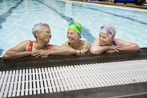 Three senior women smiling in an outdoor pool. All three are resting their arms on the pool ledge and have goggles resting on the top of their heads.