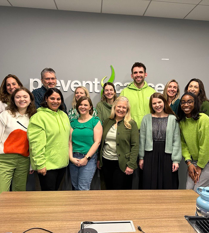 A candid photo of staff standing in front of a large Prevent Cancer Foundation logo that is mounted on a gray wall. Everyone is smiling and wearing green clothing.