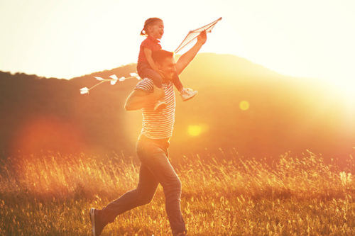 An adult man is carrying a small child on his shoulders and they are both laughing. They are moving quickly through a field in the glow of a setting sun. The man is holding up a kite with his right arm. You can see rolling hills in the background.