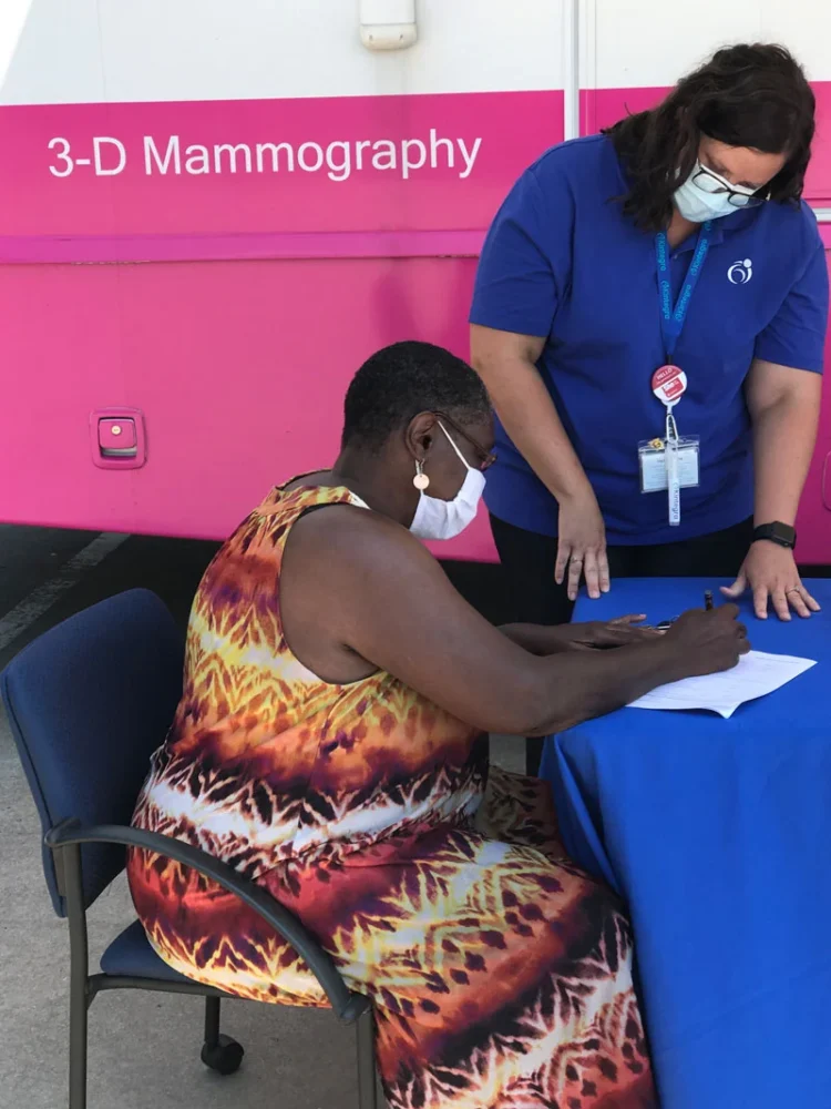 A Black woman with short hair is seated at a table outside of a bright pink mobile mammogram van. She is wearing a colorful dress, a face mask and is filling out paperwork. A clinic nurse is standing next to to assist.