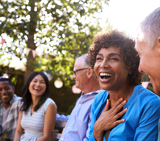 A group of older multi-racial adults are seated outside in a half-circle engaged in joyful conversation. The camera is focused on a laughing Black woman who is placing her right hand across her chest and laughing.