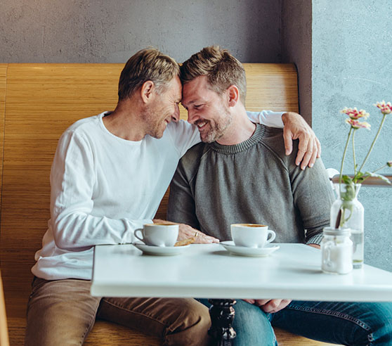Two white males in their 40s or 50s are seated at a cafe table. One man has his arm around the other and they are leaning into each other with foreheads touching. They are smiling and seem to be sharing an intimate moment.