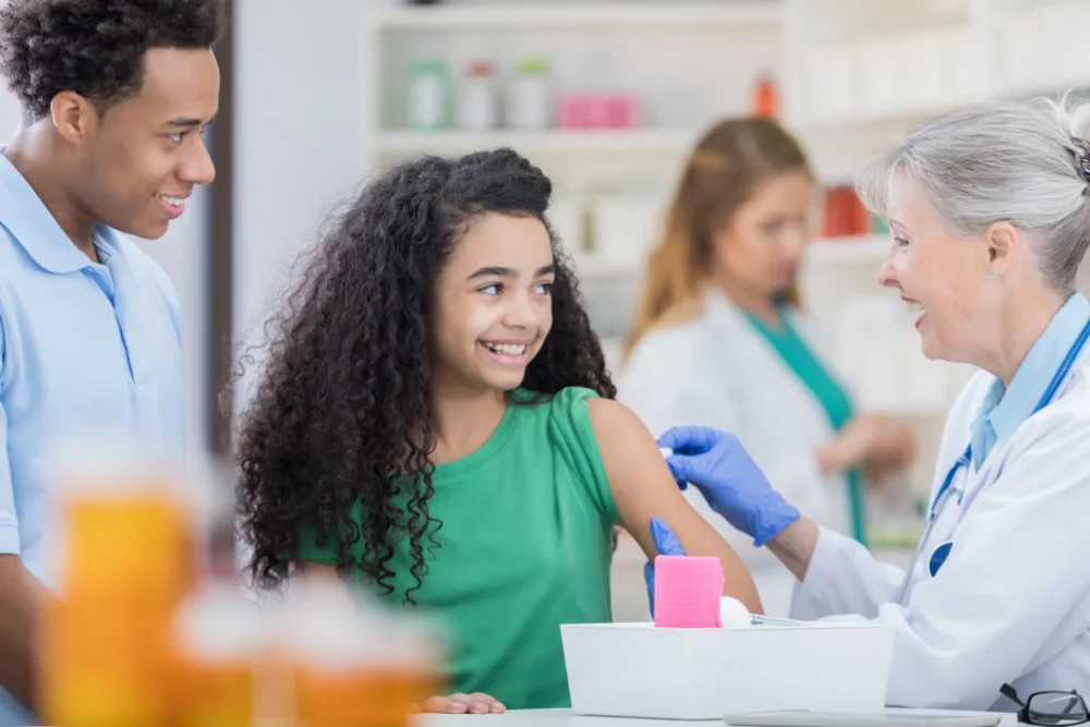 Health care professional rubs alcohol on cheerful tween girl's arm. The girl is preparing to receive back to school vaccines. Her father is smiling and standing next to her.