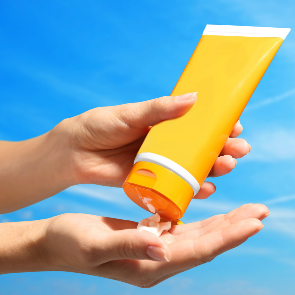 One hand squirts an orange bottle of sunscreen into another hand. In the background, the sky is blue and sunny.
