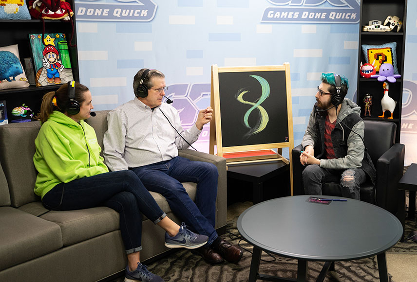 An adult woman and man with headsets sit on a couch on a stage across from another man also wearing a headset. There is a chalkboard easel between them with a green double helix drawn on it. The set background has the Games Done Quick logo repeated. There are various video game memorabilia placed on black shelves on both sides of the stage