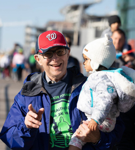 A three-quarter view of an older white man who is smiling and pointing to the camera. He is holding a toddler who is dressed in a winter jacket and white stocking hat. The man is also wearing a jacket and a navy blue t-shirt with a bright green Prevent Cancer Foundation imprint.