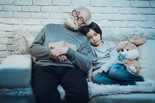 A grandpa is sitting on a couch with his grandchild at night. They both have their eyes closed and resting their heads on each other’s shoulders. The grandpa is holding a book and wearing reading glasses. A stuffed bunny is next to the little boy.