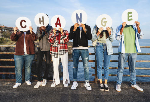 Multiracial young people cover faces with "CHANGE" discs by a railing, sea, and rocks, symbolizing a call for societal transformation