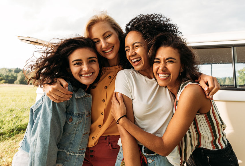 Four young women of different ethnicities gather with their arms around each other’s shoulders. They appear to be outdoors with the wind blowing their long hair. All four are laughing.