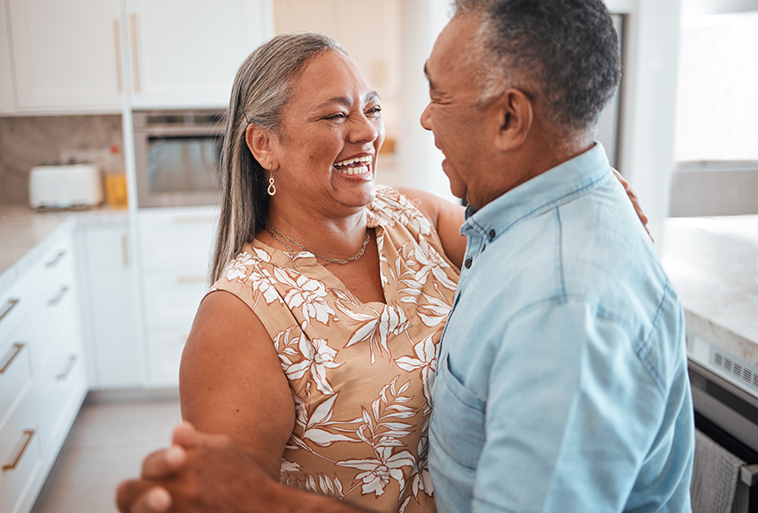 A senior man and woman dance closely in a well-lit kitchen. they are gazing at one another and smiling.