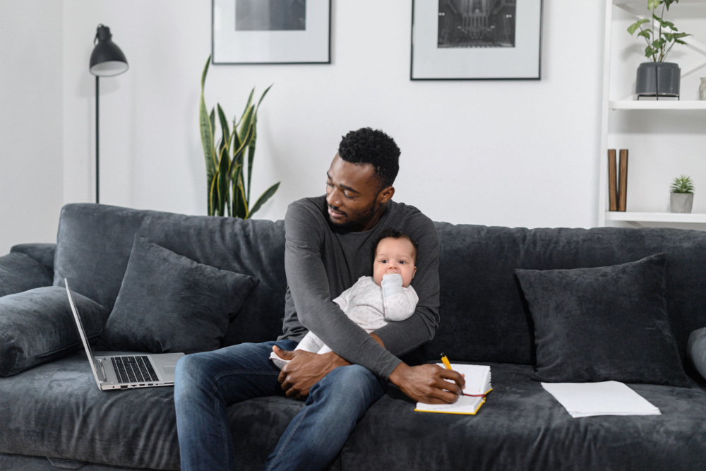 A man in his 30s or 40s sits on a couch with a baby in his lap. He is looking at a computer screen and writing down a note with his free hand.