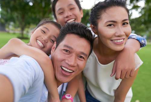 A young family of southeast Asian decent are gathered closely together in a park. The mother and father are in the front while the two school-aged children hug from the back.