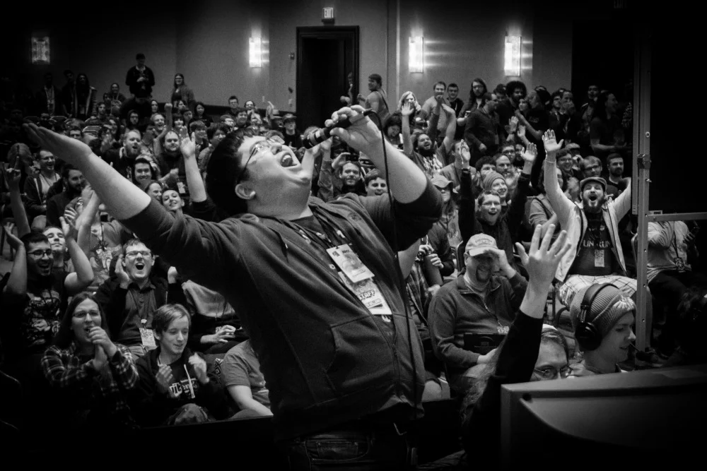 A black and white photo from the Awesome Games Done Quick event ballroom. There is a man standing in front, holding a microphone and appears to be speaking excitedly. There are dozens are people behind them celebrating and clapping.