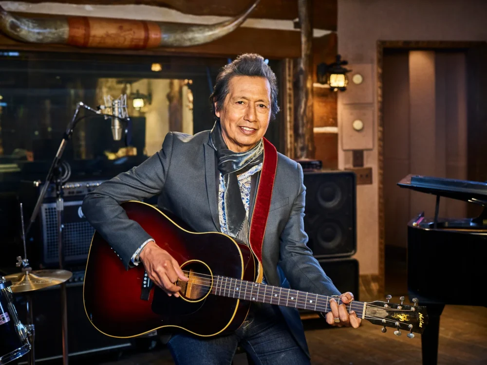 A three-quarter view of musician Alegandro Excovedo in a recording studio. He is an older Latino man seated and holding an acoustic guitar. He is wearing a gray blazer, blue jeans and a gray scarf. He is strumming the guitar and smiling at the camera.