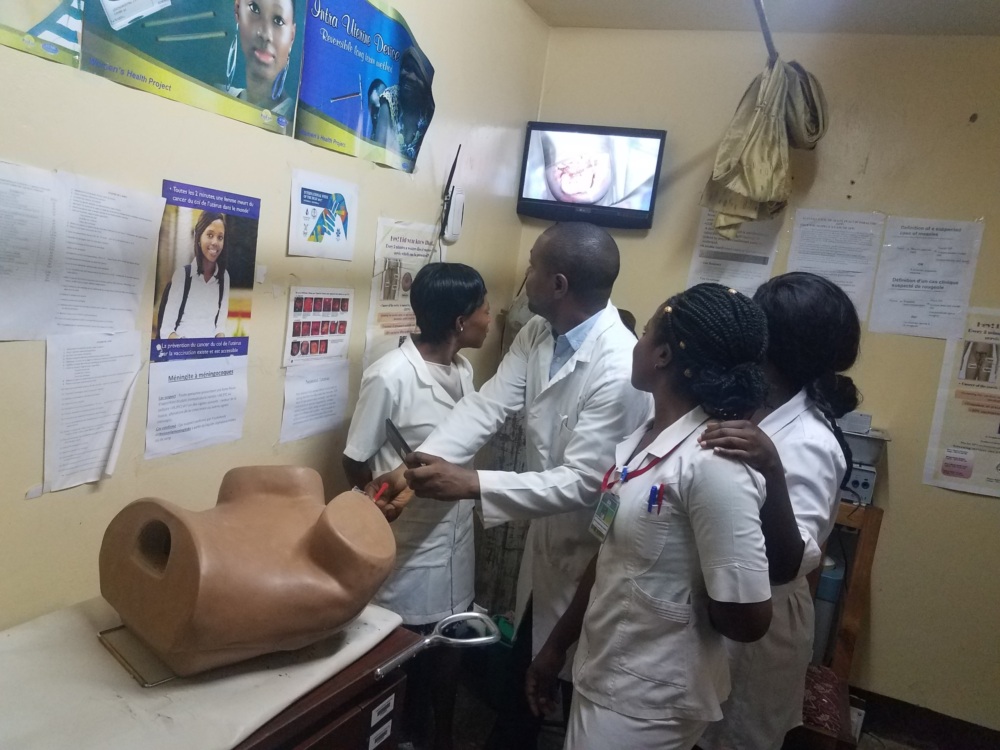 A group of four health care providers in Cameroon practice a medical procedure on a plastic model of a body. They look up at a screen with a simulation of the procedure.