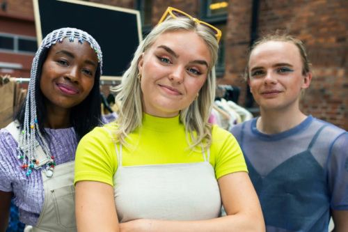 Three people stand together, dressed in bright clothes, looking confident and facing the camera.