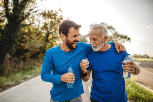 Two men exercising outdoors, walking together and laughing while one holds a water bottle.