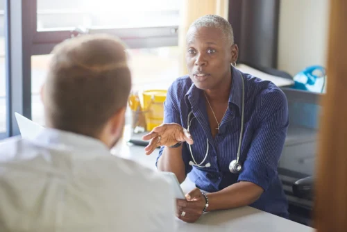 A female doctor sits at her desk and chats to a male patient while looking at his test results on her digital tablet. She is wearing a blue shirt with the sleeves rolled up and a stethoscope around her neck.