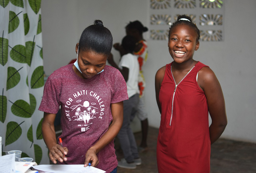 A young girl is waiting to get her HPV vaccination in Haiti. She is standing next to her mother who is looking down at paperwork. The girl is wearing a red dress, has her hair back in a bun and is smiling brightly at the camera.