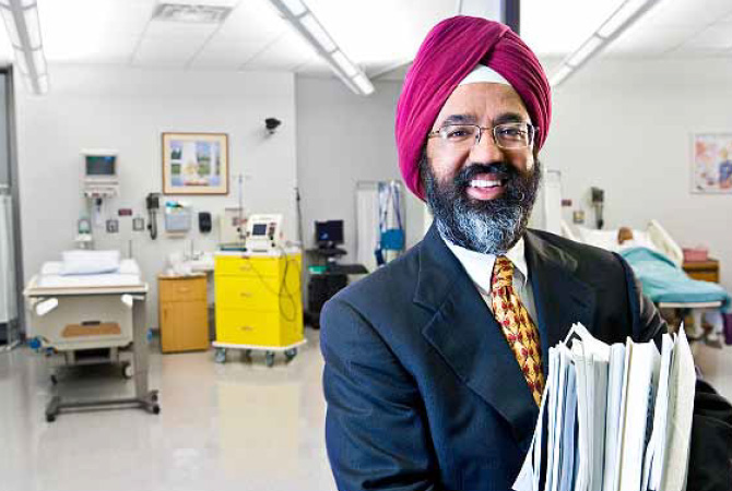 Portrait of Jasjit Ahluwalia, an Indian man wearing a turban and standing in a hospital. He has a graying beard, wearing glasses and is grinning. He is wearing a suit and holding a large stack of papers.