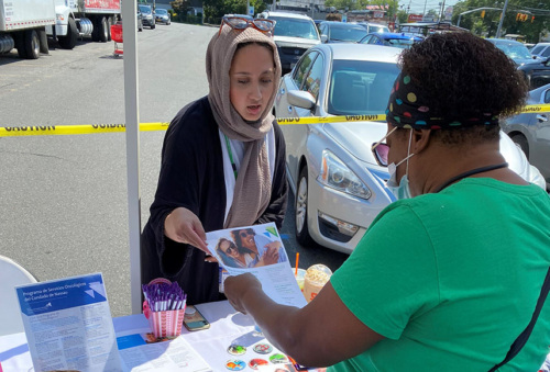 A woman is standing behind an informational table handing materials to another woman. They are outside in a parking lot.