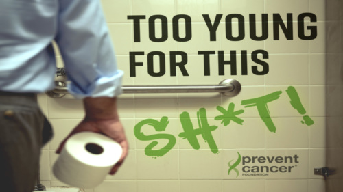 A man in his 40s wearing dress clothes is facing away from the camera in front of a toilet in a stall, holding a roll of toilet paper in his right hand. There is text painted on the wall tiles next to the toilet that reads, “Too Young for This Sh*t!”