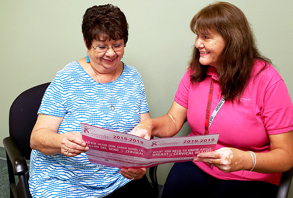 Two women in their 40s or 50s sit in a New Mexico clinic next to each other. The woman on the right is sharing an informational brochure about breast and cervical cancer screenings. The brochure is in both English and Spanish.