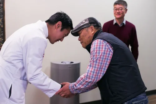 A doctor of Korean descent bows and shakes hands with an elderly man also of Korean descent.