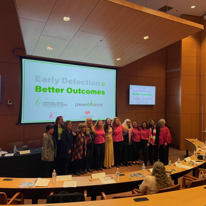 A group of 13 women pose to take a picture together in an auditorium with a sign behind them that says Early Detection = Better Outcomes.