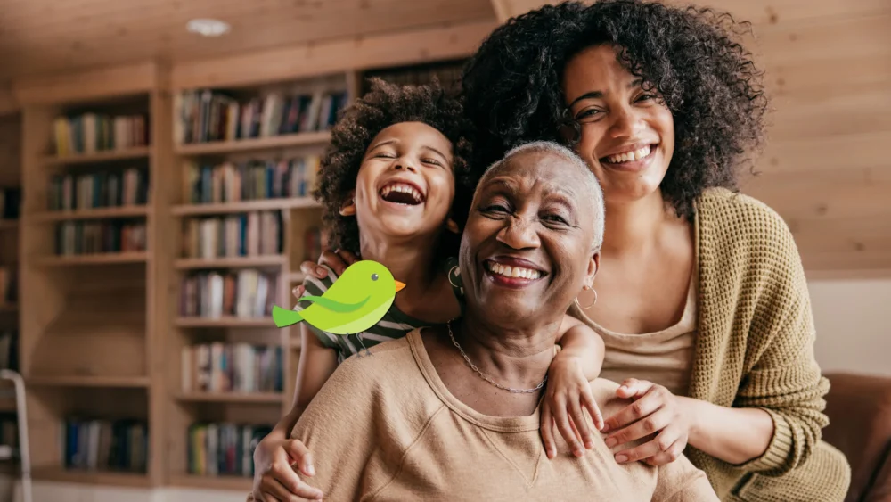 A Black grandmother is seated with her daughter and grandson hugging her from behind. All three are facing forward and grinning. There is a green cartoon bird with an orange beak resting on Grandma's shoulder.