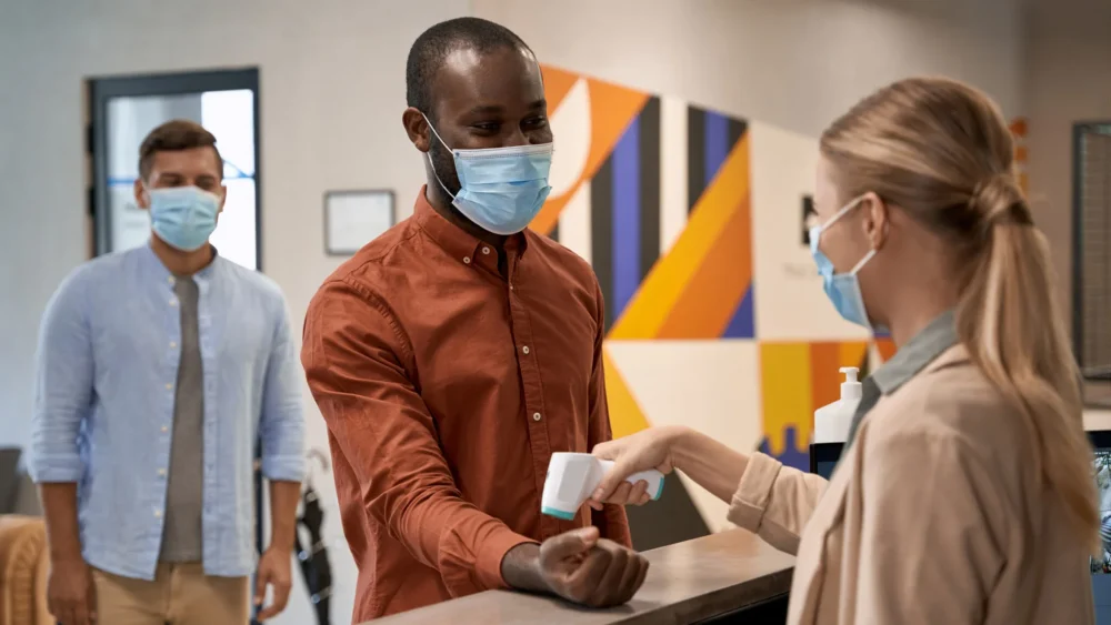 A man in his 40s, wearing a mask, having his temperature taken during a doctor’s visit.