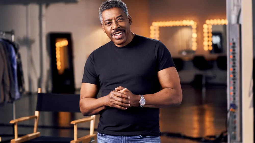 Three-quarter view of actor Ernie Hudson. He is an older Black man wearing a black t-shirt and jeans. He is clasping his hands together and smiling at the camera.