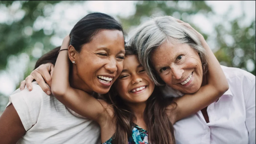 A girl with brown skin and hair is between her mother and grandmother facing forward. The girl is spreading her arms and wrapping them around the heads of her mother and grandmother in a hug.