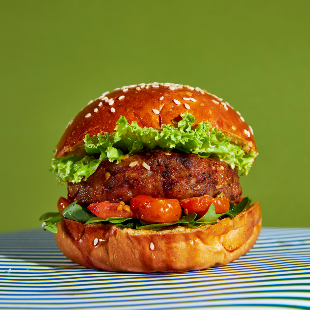 A veggie burger rests on a table with a green and white striped tablecloth. The burger contains a veggie patty, tomatoes and greens sandwiched between a burger bun with sesame seeds. The background is green.
