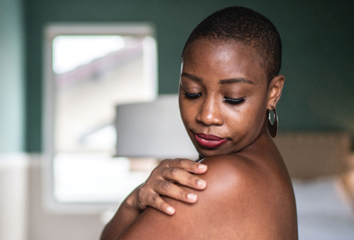 A three-quarter portrait of a black woman in her 40s who is wearing just a towel. Her right arm is wrapped around her front and touching her left shoulder. She is looking down at her shoulder's skin.