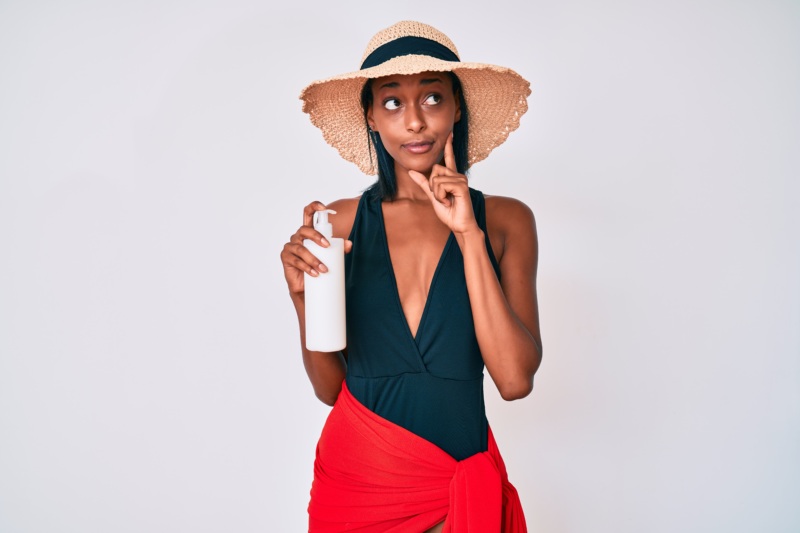 A Black woman in a sunhat puts her finger up to her face in an inquisitive look. In her other hand she holds a bottle of sunscreen.