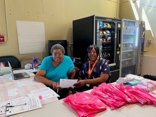 Two women sit at a table at the Research Foundation for The SUNY of Univ. at Buffalo. Plastic bags and forms are on the table, while vending machines are behind them.