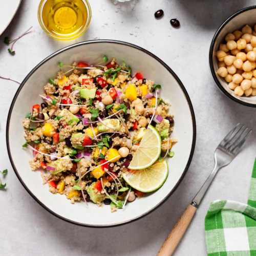A bow has lime slices, quinoa, red onion, avocado an chickpeas in it. Also on the table is a green checkered napkin, a fork, a bottle of oil and a bowl of chickpeas.