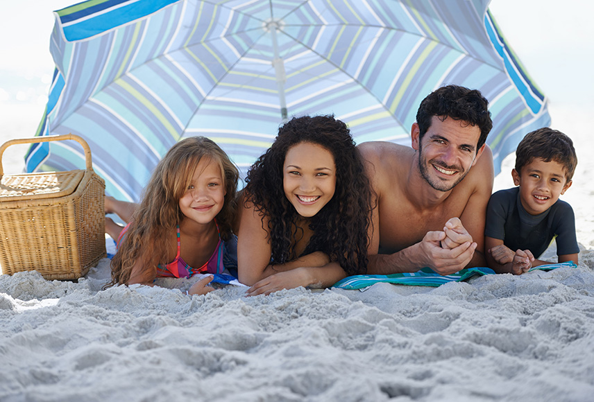 A mixed race family of four seeks shade from the sun under a beach umbrella. The family is lying side-by-side on their stomach, propped up on their elbows and smiling.