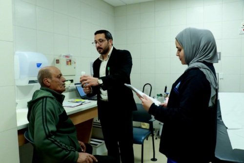 An Egyptian doctor is standing and taking the temperature of a senior man who is seated. A nurse is standing to the right and is reading the patient's chart.