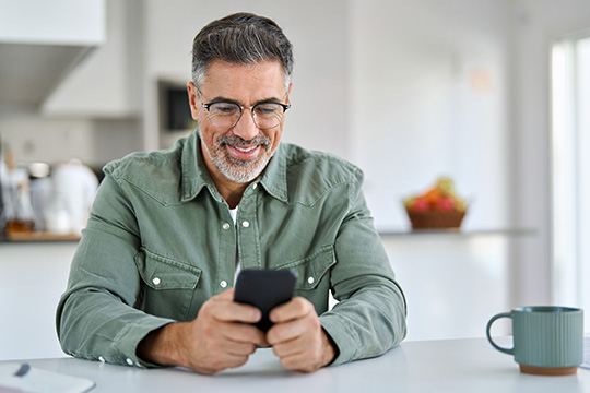 Middle-age man with dark, graying hair and beard holds a smart phone in both hands. He is wearing horn-rimmed glasses and smiling. He is seated at a kitchen table and a mug is next to him.