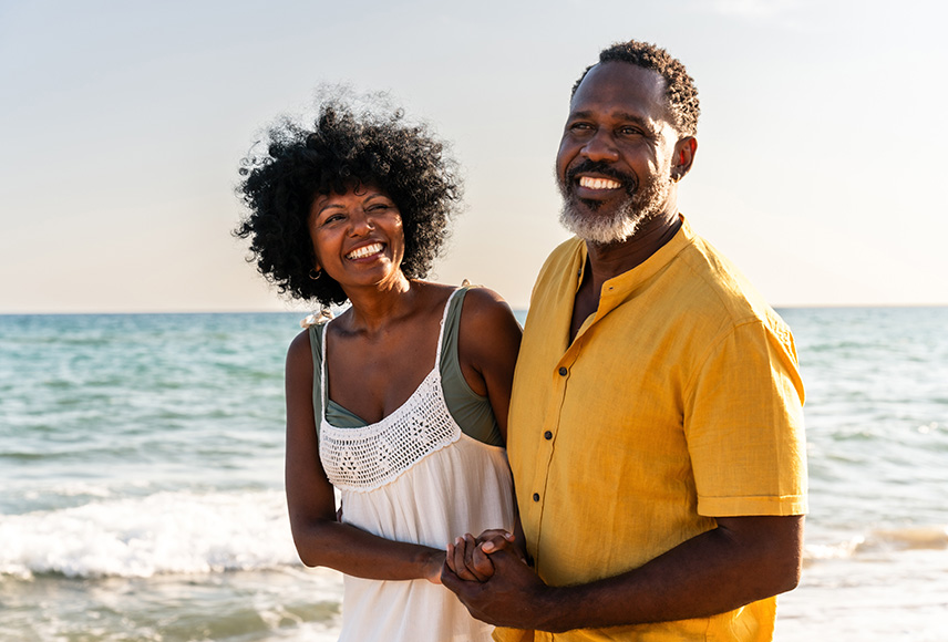 A Black woman and man in their 50s are strolling on a sunny beach. Both are smiling and they are holding hands.