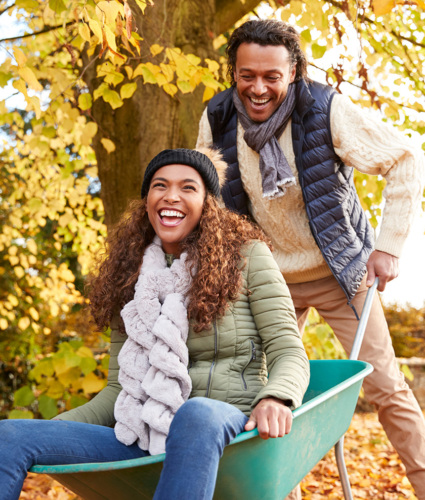 A Black woman in her 30s or 40s is seated in a wheelbarrow. She is dressed in a jacket, jeans and scar and is laughing. The wheelbarrow is being pushed by a Black man in his 30s or 40s. He is wearing a sweater, puffer vest, scarf and khaki pants. He is looking down at the woman and smiling. The leaves in the background have turned colors indicating it's fall.