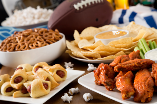 A table with pigs in a blanket, buffalo wings, potato chips, pretzels, popcorn and a football.